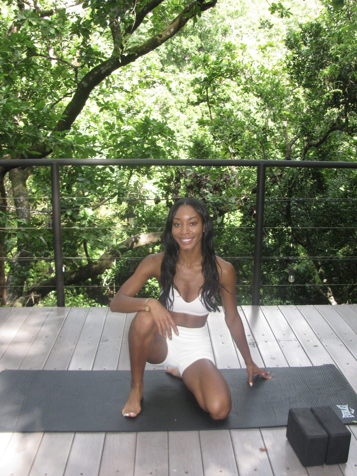 Maya smiling, kneeling on her mat on a wooden deck among the trees