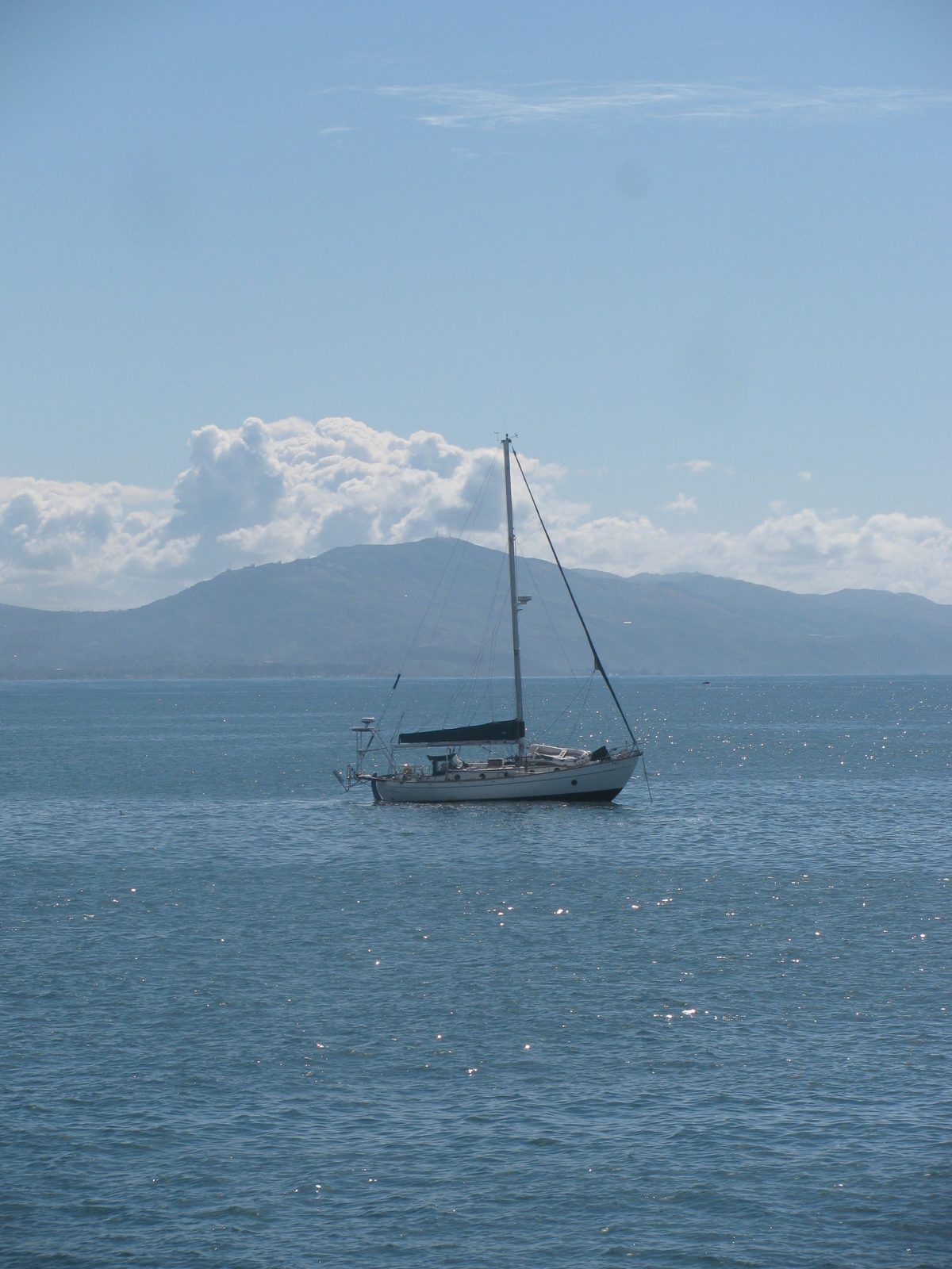 A sailboat at anchor on a glittering bay, green mountains and white clouds beyond